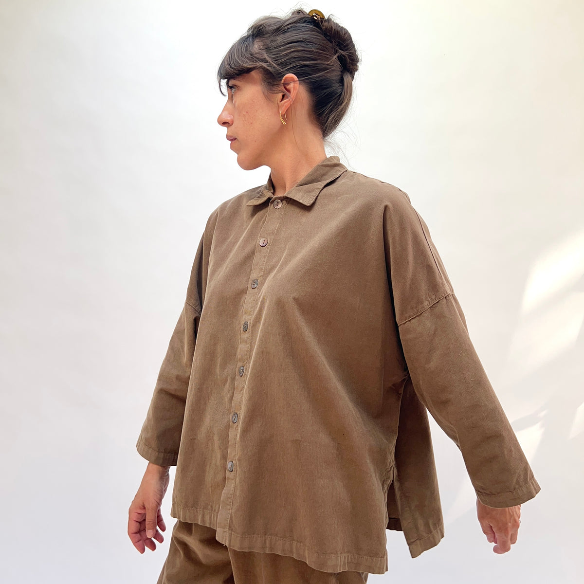 Side profile of a woman wearing the Eleven Stitch Corduroy Big Shirt in Winter Olive, showing the classic point collar, shell button front, dropped shoulder seam with topstitching, wide sleeve with banded cuff, and straight cropped hem in fine-wale olive cotton corduroy, styled with matching corduroy wide-leg trousers against a white background
