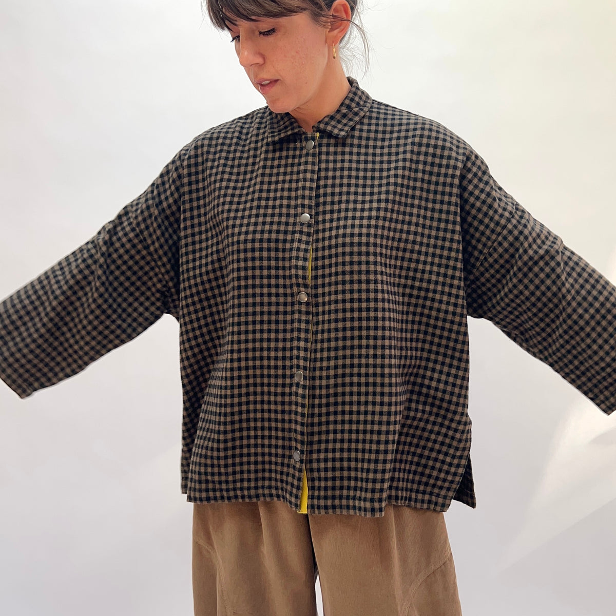 Front view of a woman wearing the Eleven Stitch Small Check Big Shirt in Winter Olive with arms outstretched, showing the point collar, full snap button front with contrast yellow topstitching, chest patch pocket, dropped shoulders, wide sleeves, and side slits at the hem in olive and black small gingham check cotton, styled with tan wide-leg trousers against a white background
