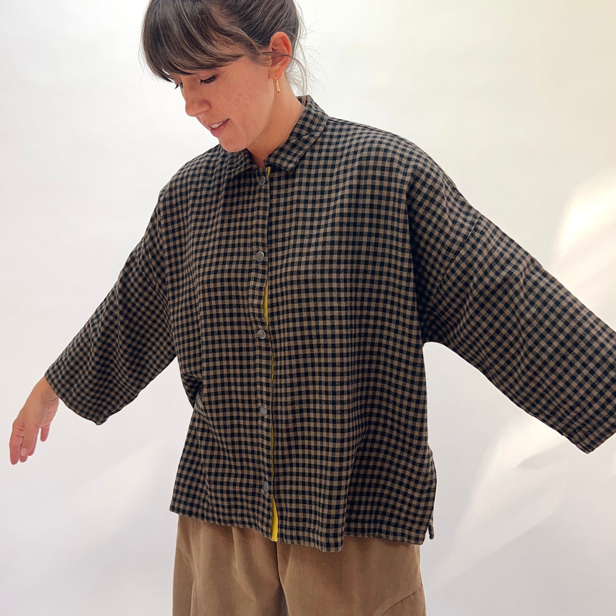 Three-quarter front view of a woman wearing the Eleven Stitch Small Check Big Shirt in Winter Olive looking downward, showing the point collar, snap button front with contrast yellow topstitching, chest patch pocket, dropped shoulders, wide 3/4-length sleeves, and straight cropped hem with side slits in olive and black small gingham check cotton, styled with tan wide-leg trousers against a white background

