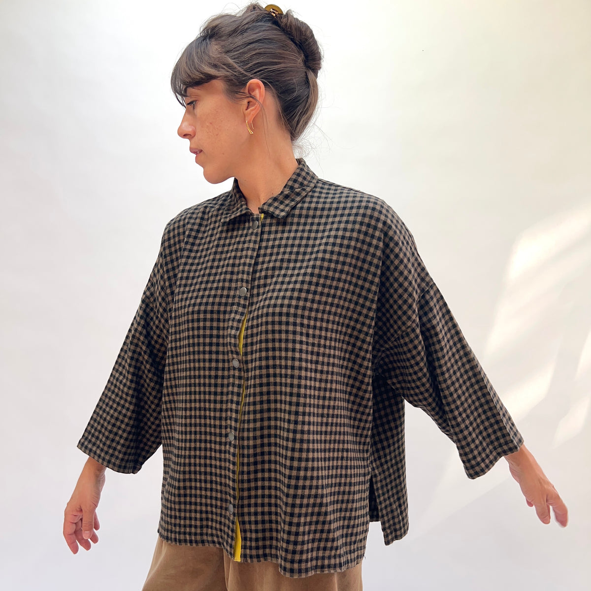 Three-quarter side view of a woman wearing the Eleven Stitch Small Check Big Shirt in Winter Olive, showing the point collar, snap button front with contrast yellow center topstitching, chest patch pocket, dropped shoulder, wide 3/4-length sleeve, and side slit hem in olive and black small gingham check cotton, styled with tan wide-leg trousers against a white background
