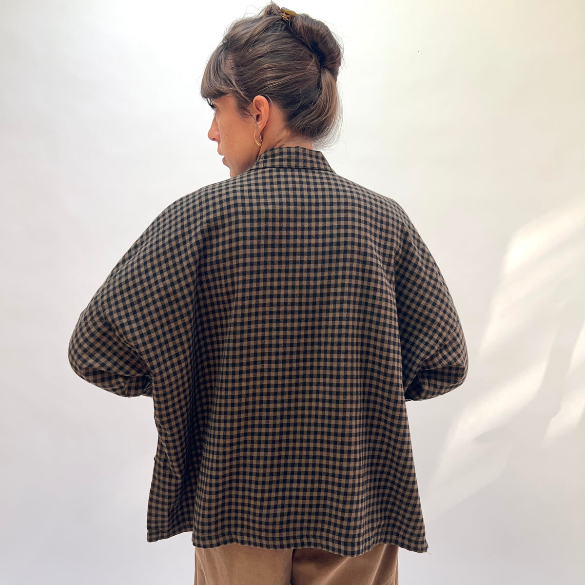 Back view of a woman wearing the Eleven Stitch Small Check Big Shirt in Winter Olive, showing the point collar, wide dropped shoulders, long sleeves, straight cropped back hem, and the boxy oversized silhouette in olive and black small gingham check cotton, styled with tan wide-leg trousers against a white background
