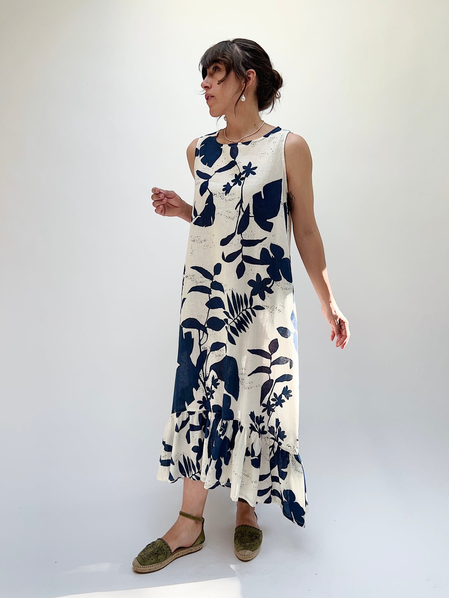 A woman models a sleeveless, knee-length dress with a foliage-inspired print. The dress features a ruffled hi-low hem.