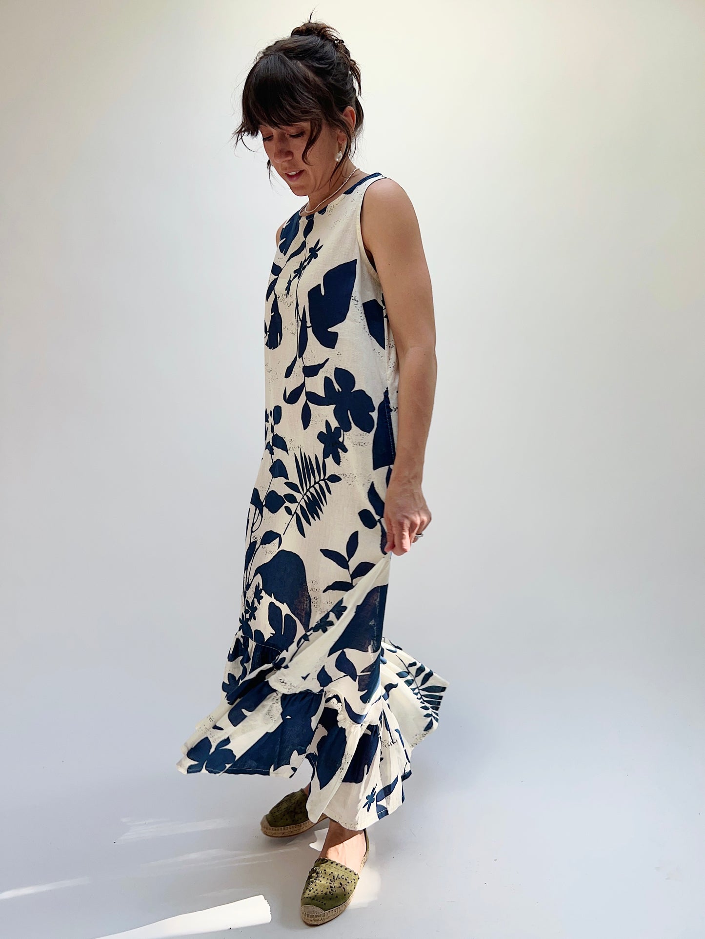 A woman models a sleeveless, knee-length dress with a foliage-inspired print. The dress features a ruffled hi-low hem.