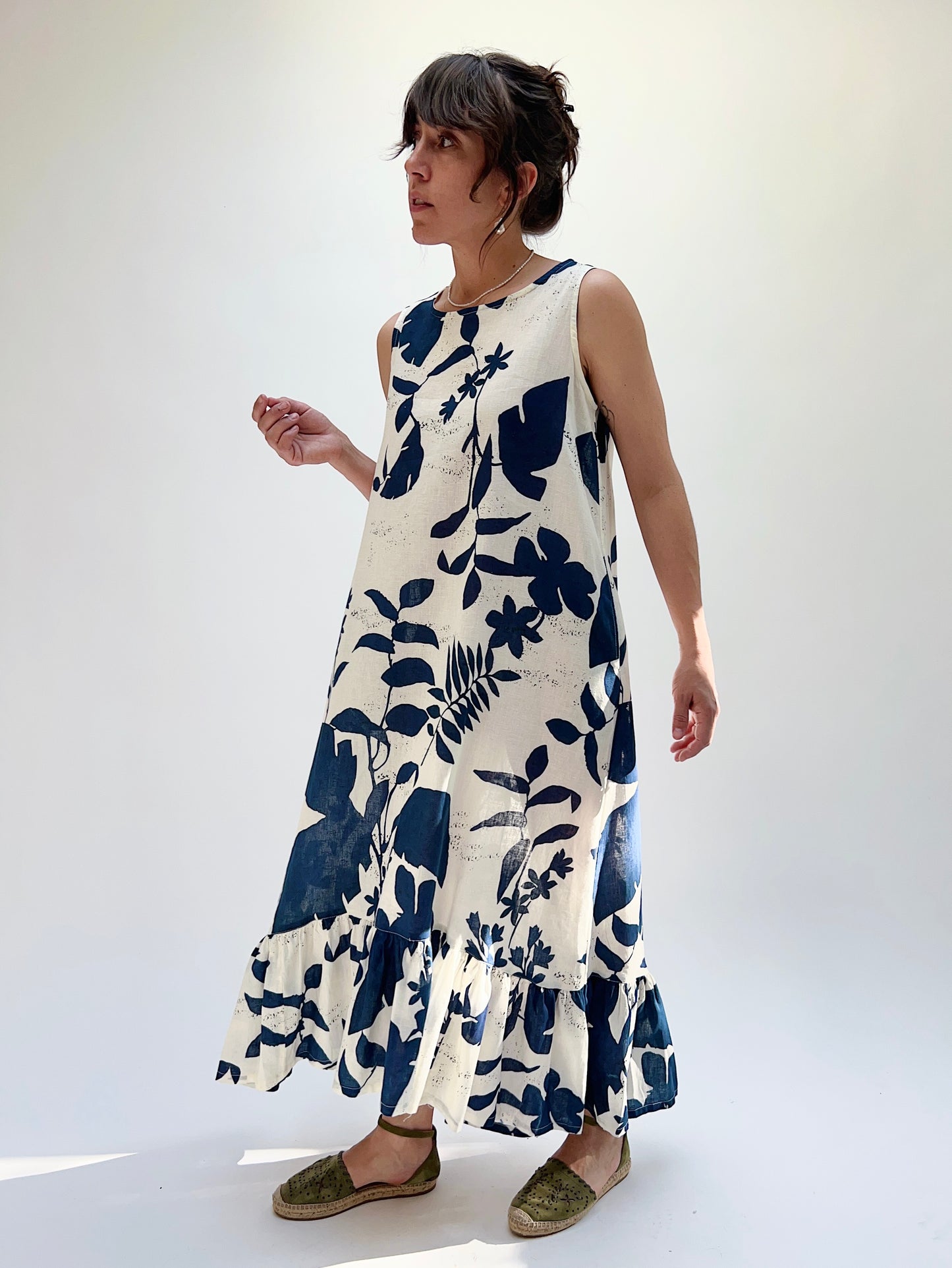 A woman models a sleeveless, knee-length dress with a foliage-inspired print. The dress features a ruffled hi-low hem.