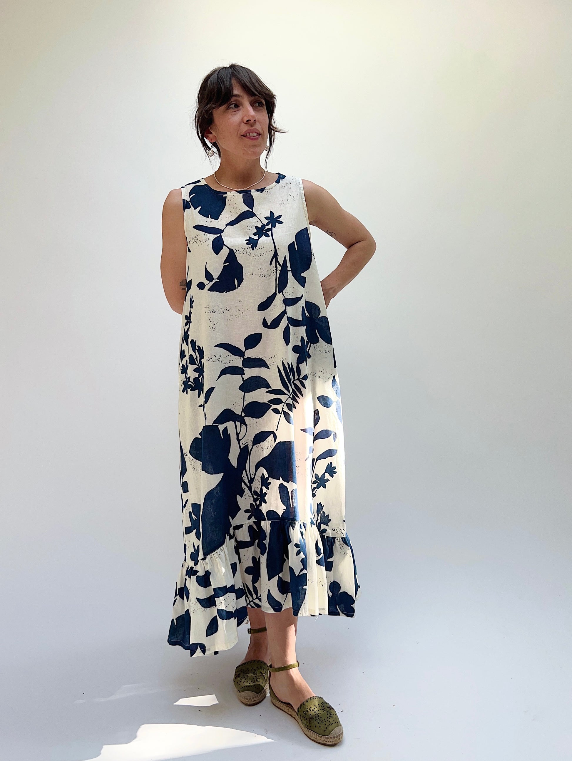 A woman models a sleeveless, knee-length dress with a foliage-inspired print. The dress features a ruffled hi-low hem.