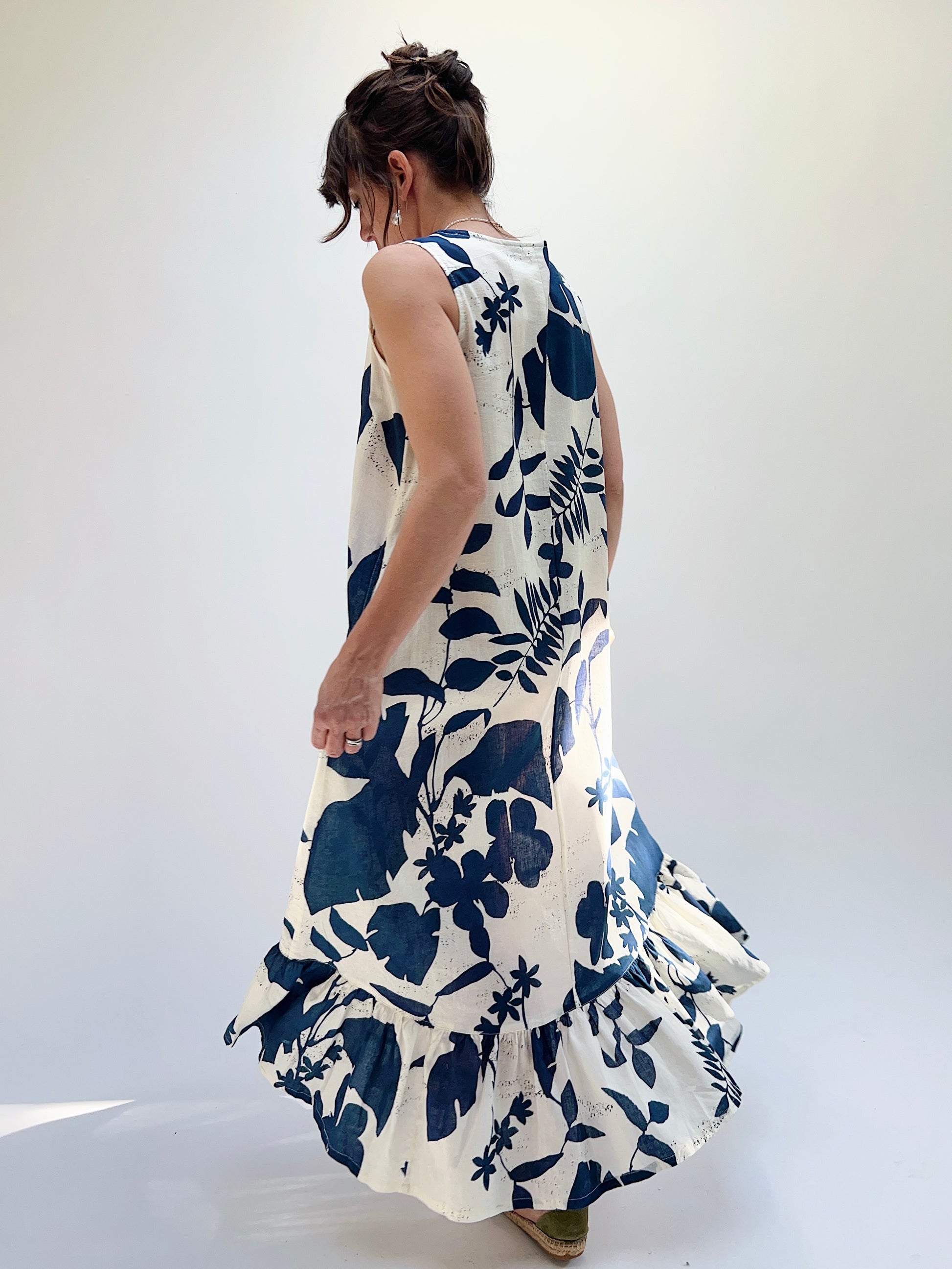 A woman models a sleeveless, knee-length dress with a foliage-inspired print. The dress features a ruffled hi-low hem, viewed from the back
