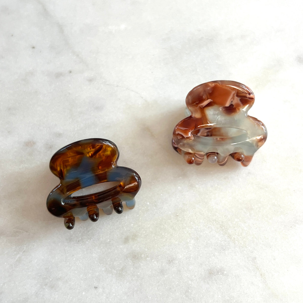 Two tortoiseshell hair claw clips on a white marble surface, one solid dark brown and one marbled with brown, black, and white.
