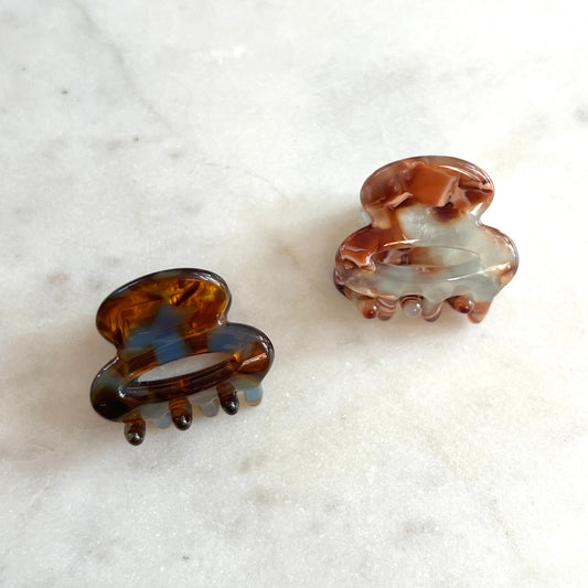 Two tortoiseshell hair claw clips on a white marble surface, one solid dark brown and one marbled with brown, black, and white.