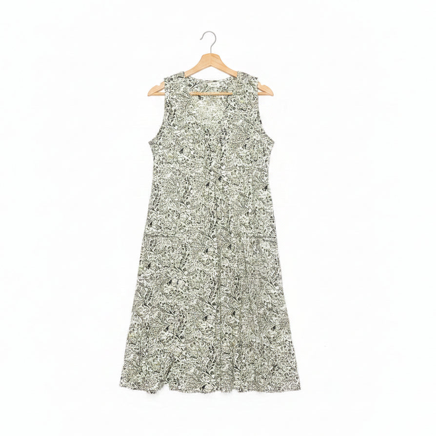 A sleeveless A-line dress with a dense green floral pattern on a white base, hanging on a wooden hanger on a dark concrete floor.