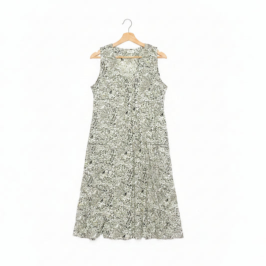 A sleeveless A-line dress with a dense green floral pattern on a white base, hanging on a wooden hanger on a dark concrete floor.