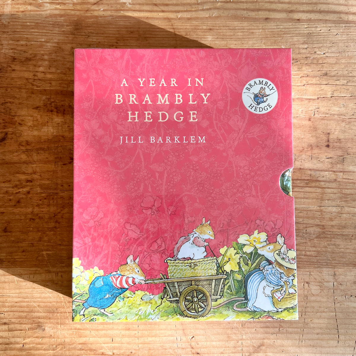 Pink slipcase for A Year in Brambly Hedge by Jill Barklem, illustrated with mice pushing a cart among spring flowers.
