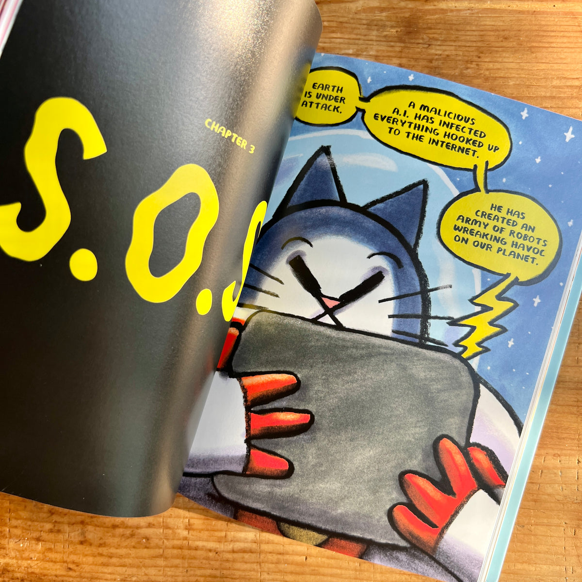 Open book showing the chapter title “S.O.S.” and a comic illustration of First Cat learning that a malicious AI is attacking Earth.
