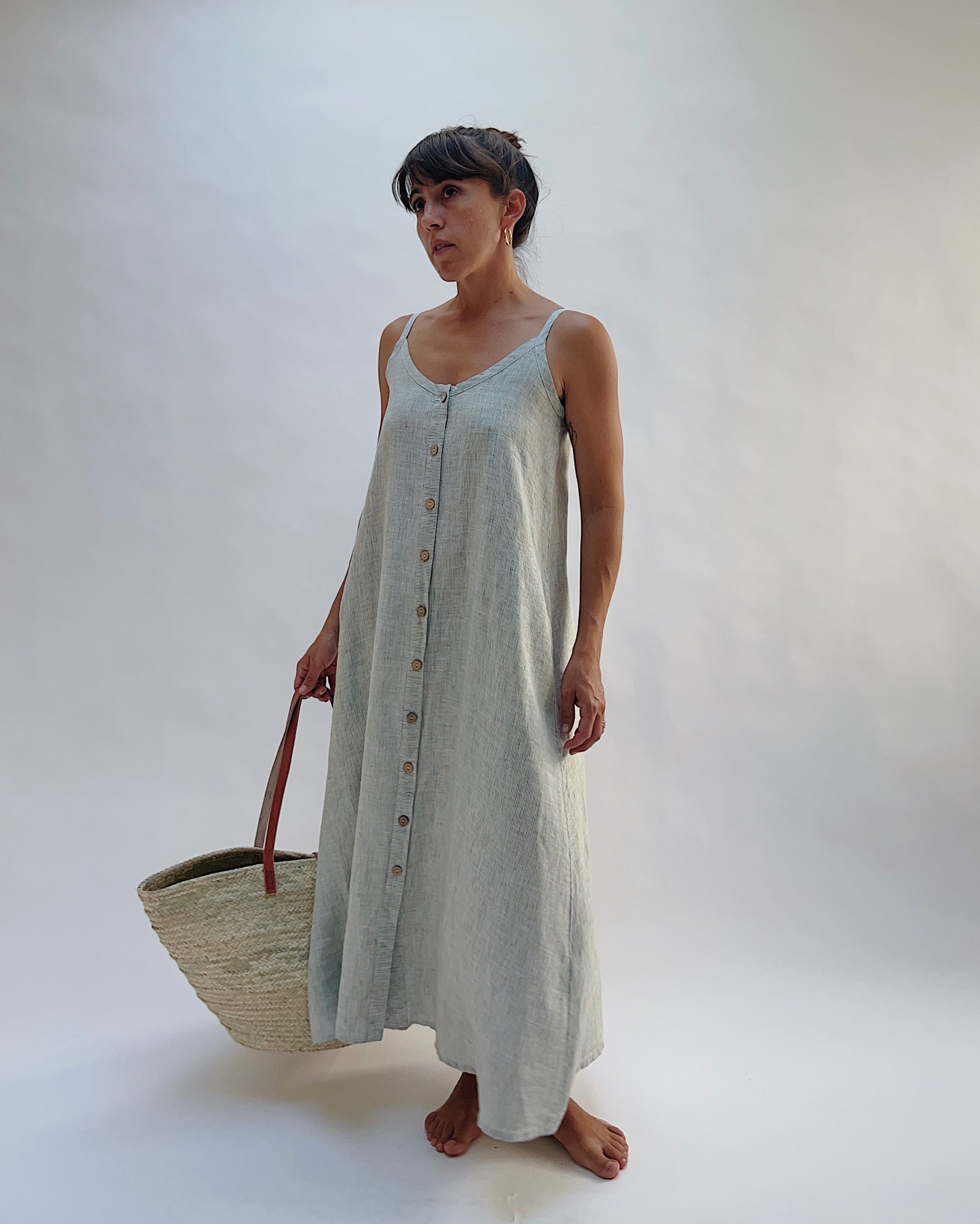 Model wearing the Yuvita Button Front Dress in Sage Pinstripe Stripe, a flowy linen maxi dress with button-front detail and spaghetti straps holding a woven tote bag