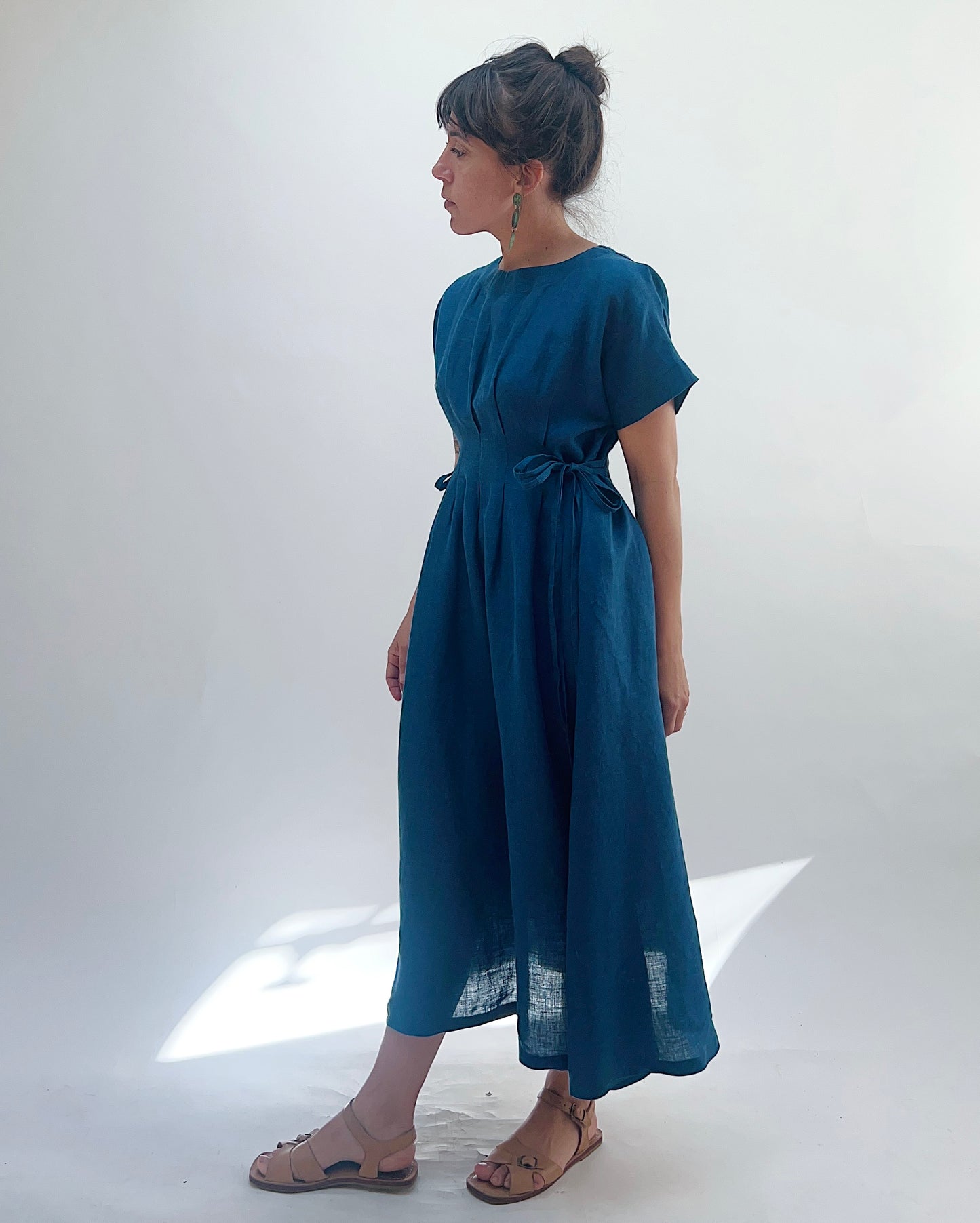 Woman with dark hair in a bun wearing a blue linen dress and green dangle earrings standing against a white background as seen from the side looking left. 