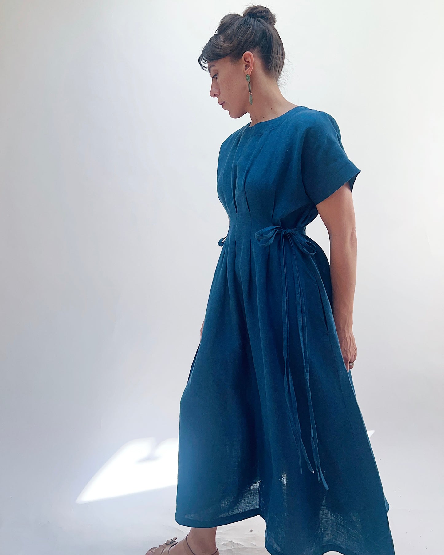 Woman with dark hair in a bun wearing a blue linen dress and green dangle earrings standing against a white background.