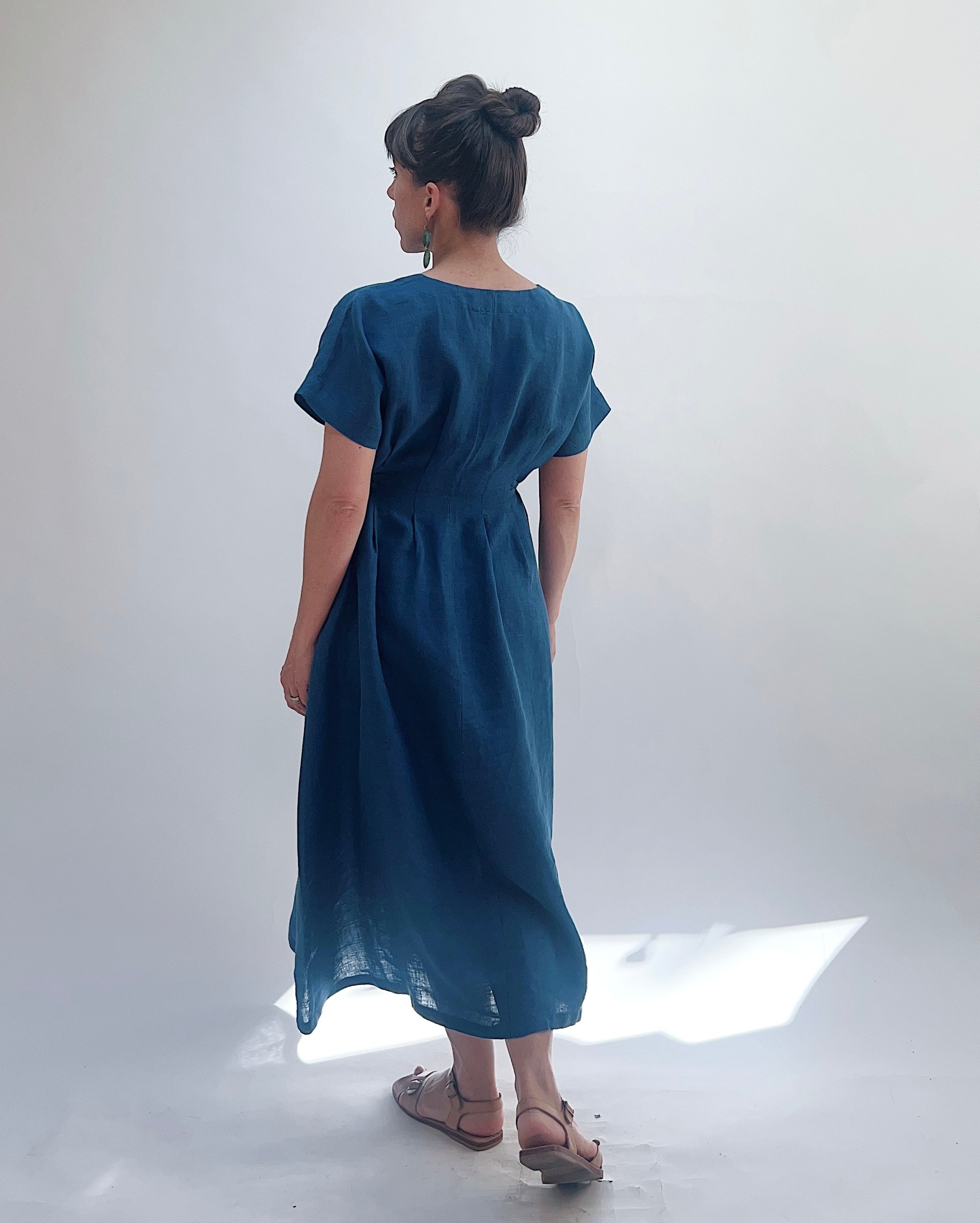 Woman with dark hair in a bun wearing a blue linen dress and green dangle earrings standing against a white background as seen from behind. 