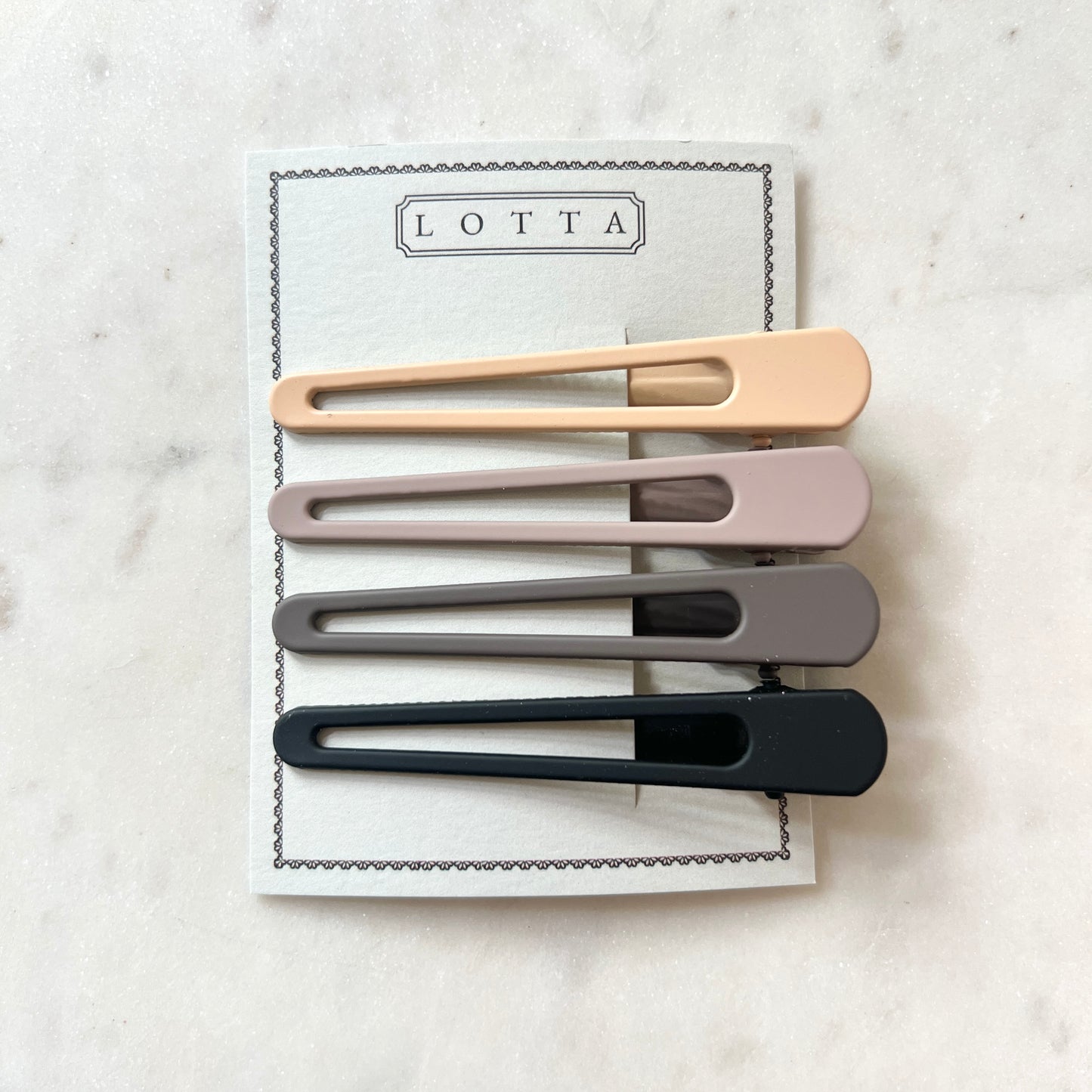 Set of four hair clips in beige, gray, and black on a Lotta branded card against a marble background.