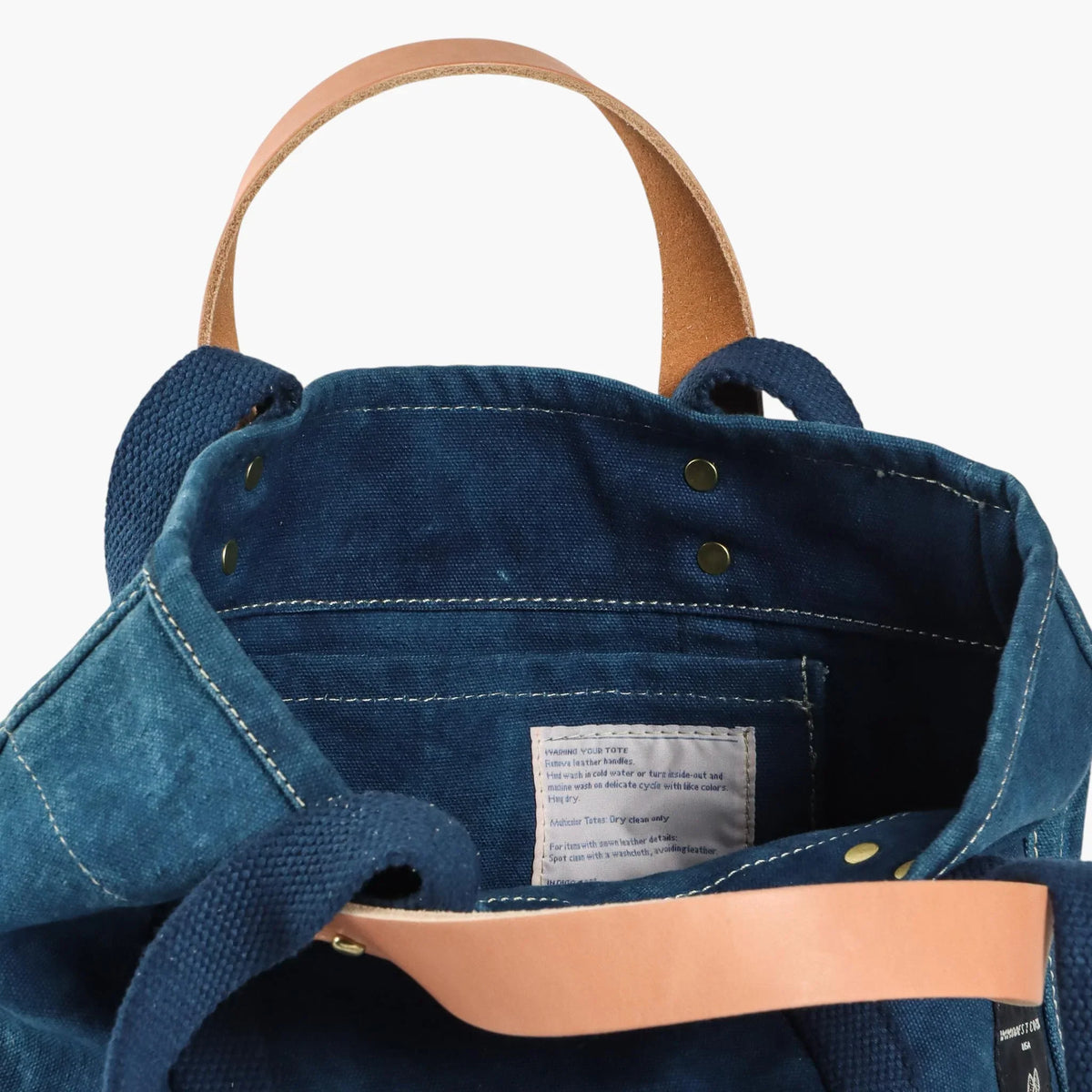 Interior of the indigo Lunch Tote showing the inside pocket, care label, and leather and webbing handles.
