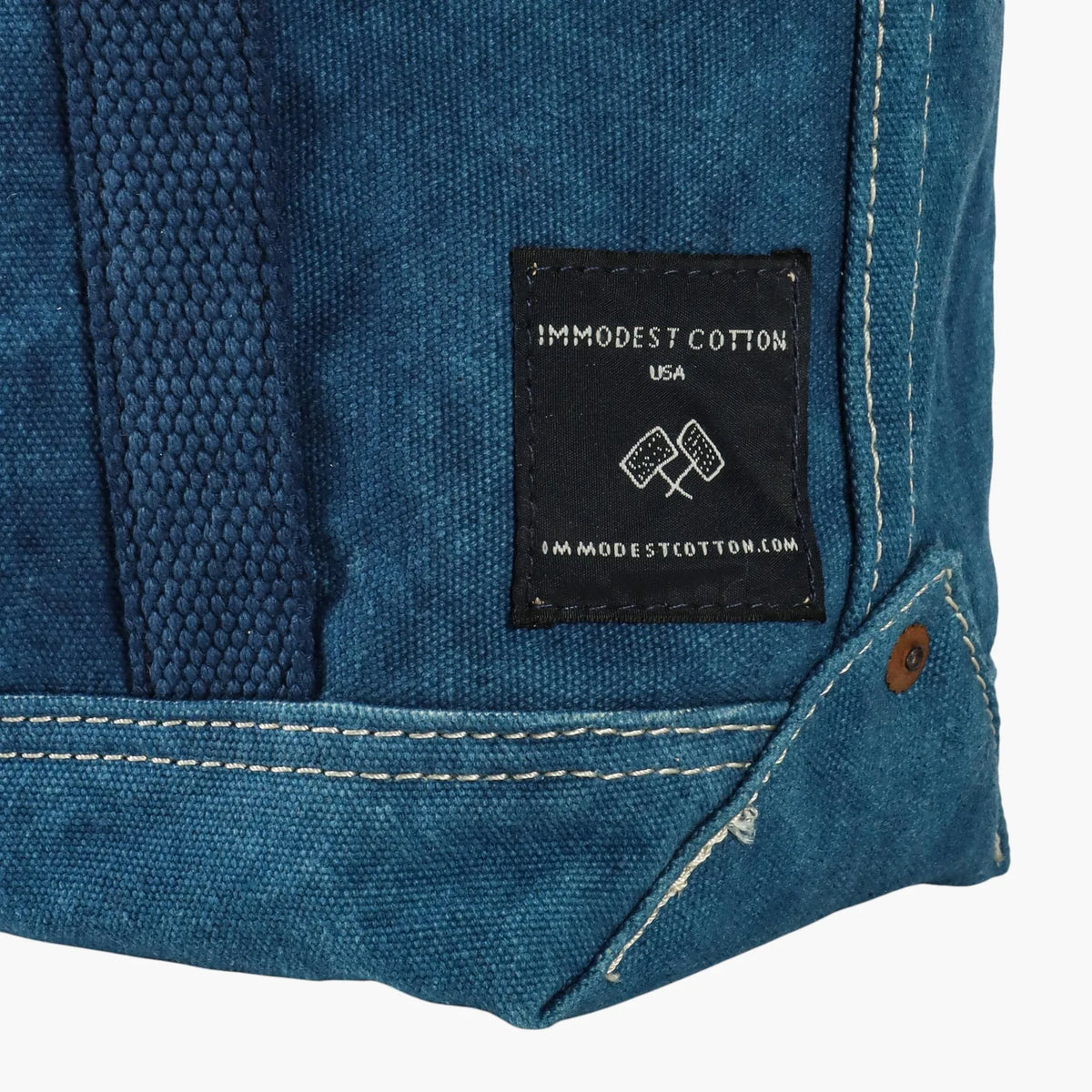 Close-up of the black Immodest Cotton label, white double stitching, and copper rivet on the indigo Lunch Tote.
