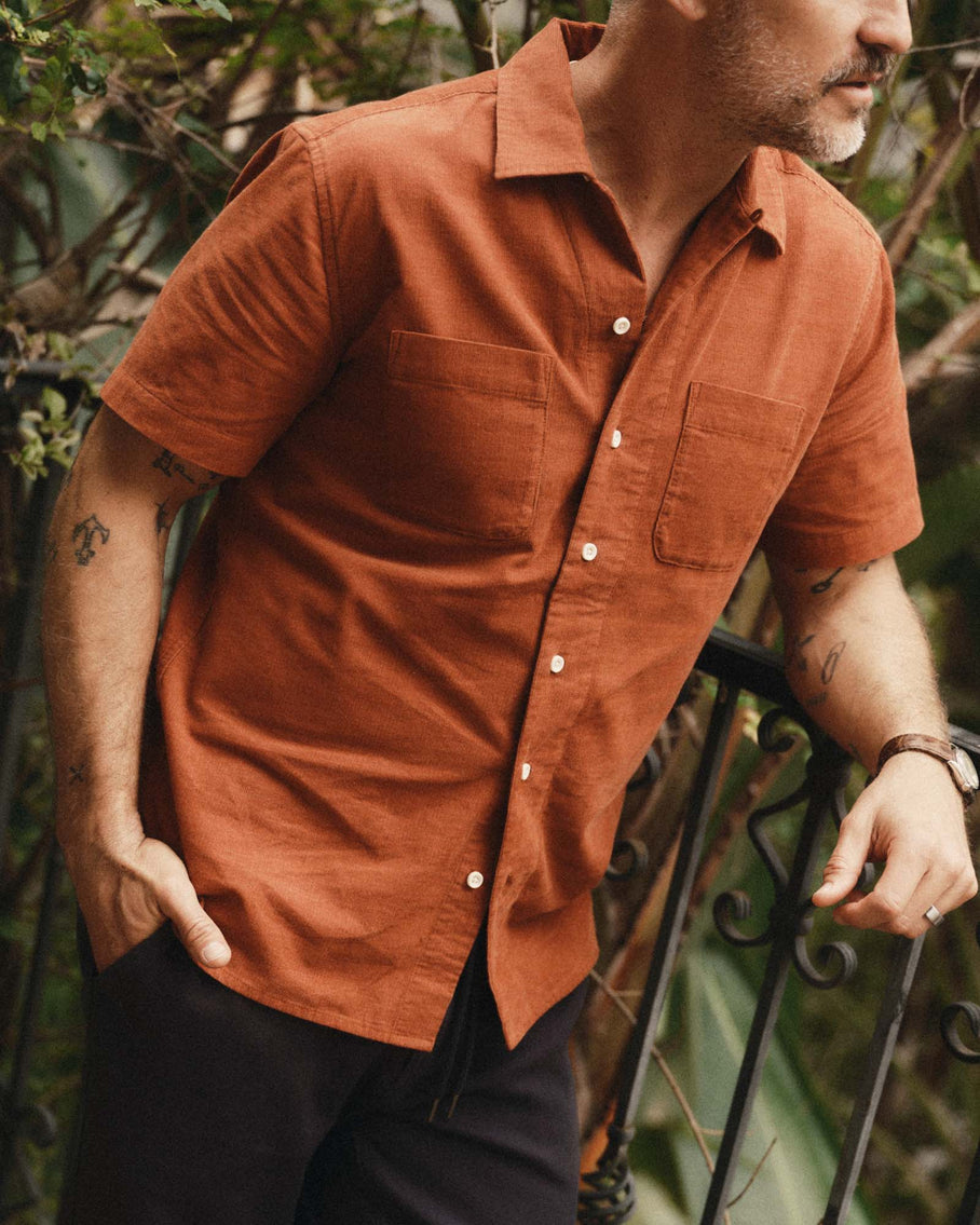 Model wearing Taylor Stitch Conrad Shirt in Heather Chili Pincord, three-quarter front view on garden balcony showing warm burnt orange organic cotton pincord, open camp collar, short sleeves, natural corozo buttons, and two rounded chest pockets, styled with dark navy trousers
