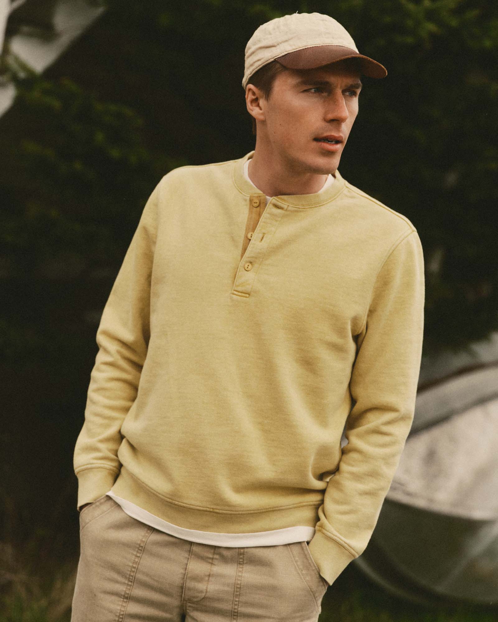 Male model wearing the Taylor Stitch Sunset Henley in washed goldfinch yellow, styled with khaki pants and a cream cap, photographed outdoors