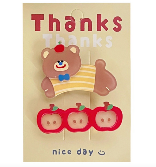 A set of two hair clips with a bear and apple design shown displayed on a paper card