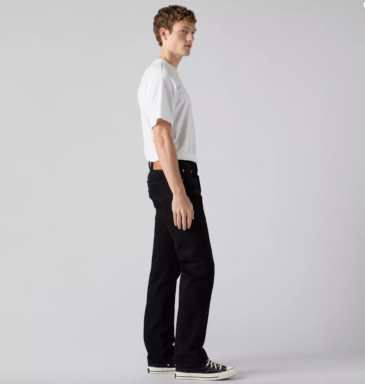 A man in a white t-shirt and black jeans is shown in profile against a light gray studio background.