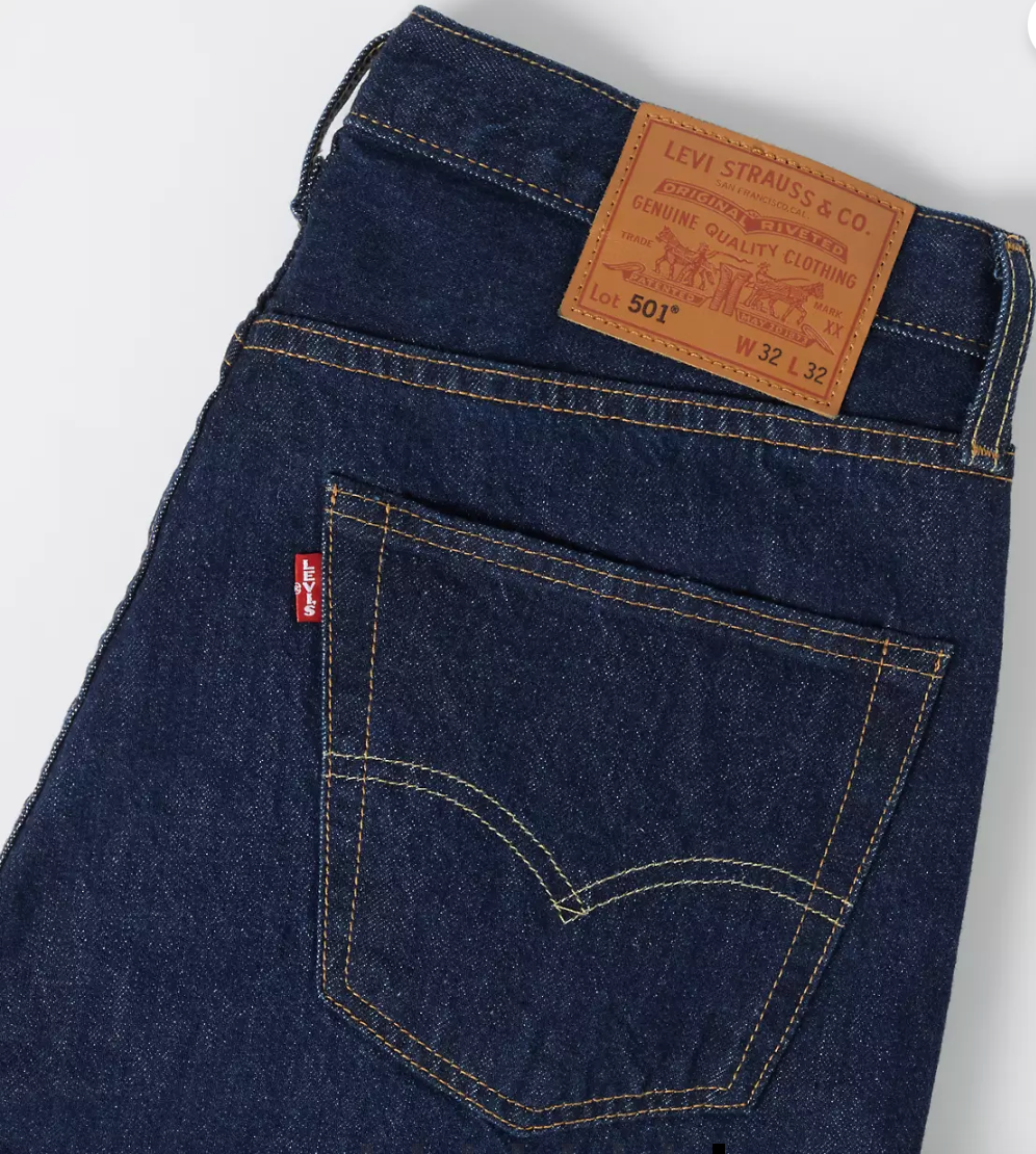 Close-up of the back waistband and pocket area of dark indigo Levi's jeans, showing the brown leather patch and red tab on a white background.
