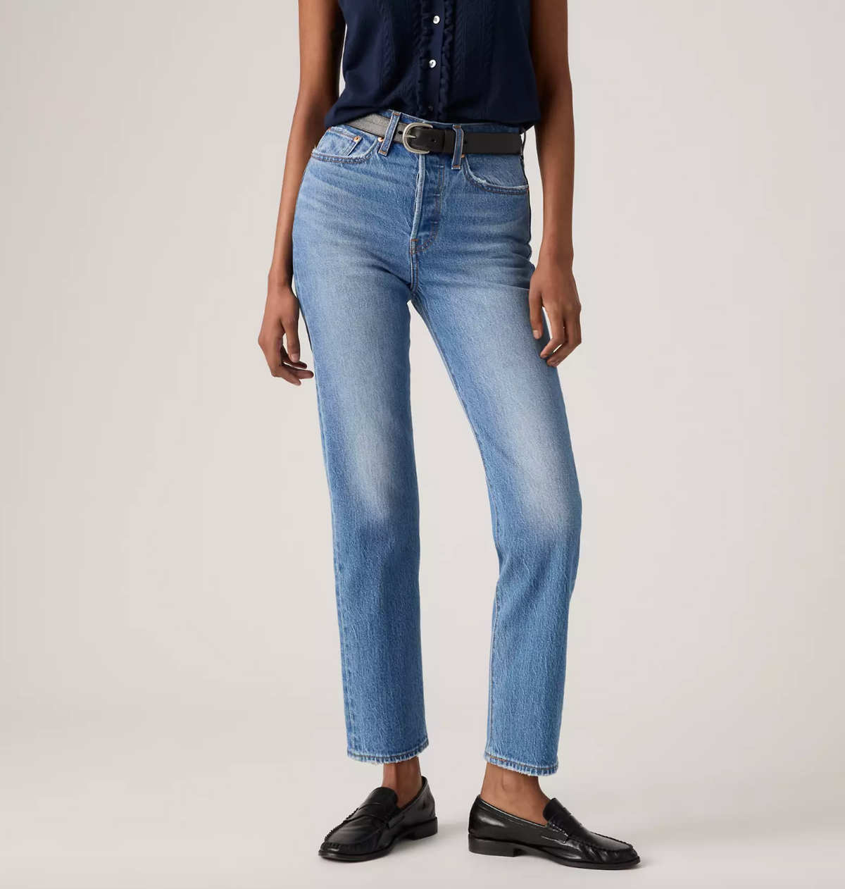 A model wearing light-wash blue straight-leg jeans with a dark belt, paired with a navy top and black loafers, photographed against a neutral light gray studio background.