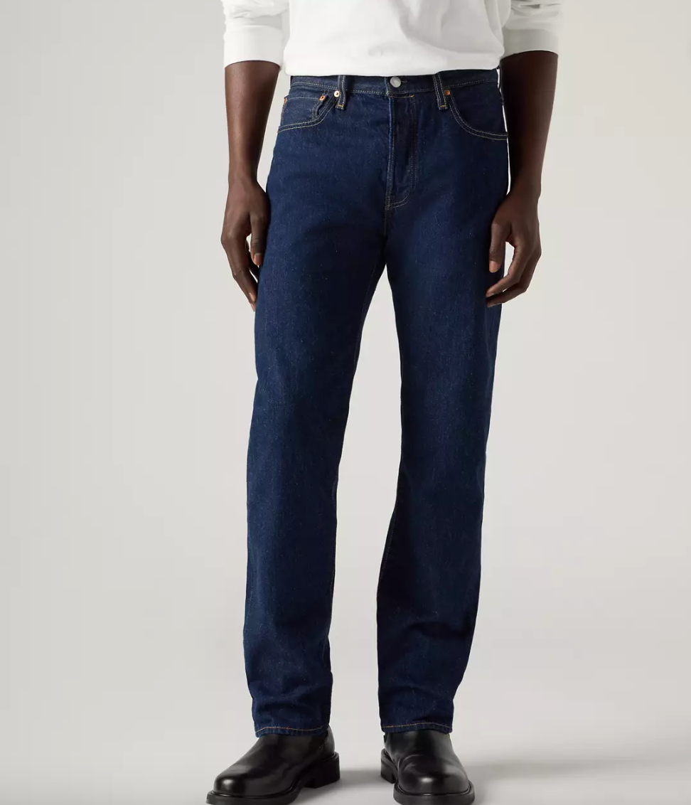 A person wearing dark blue straight-leg denim jeans, a white long-sleeve shirt, and black leather boots, photographed against a clean light gray studio background.