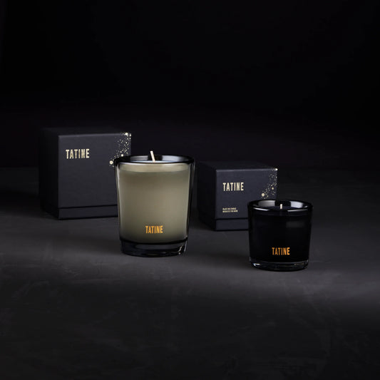 Two Tatine candles, one green and one black, with boxes in the background on a dark surface.