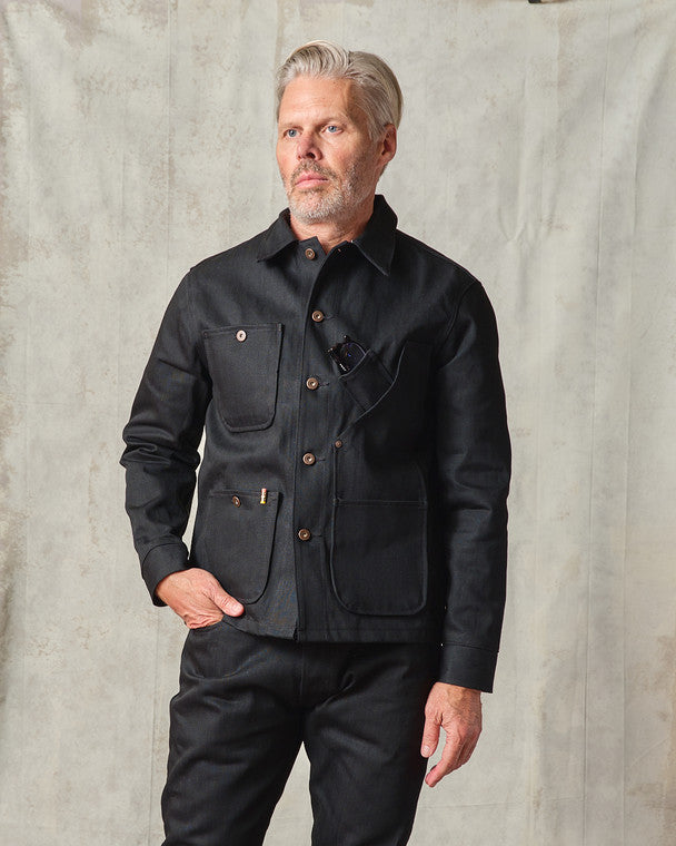 Front view of model wearing the Tellason Coverall Jacket in Black Selvedge Denim with hand in pocket, showing the full button front, chest pocket, and lower utility pockets with Tellason label