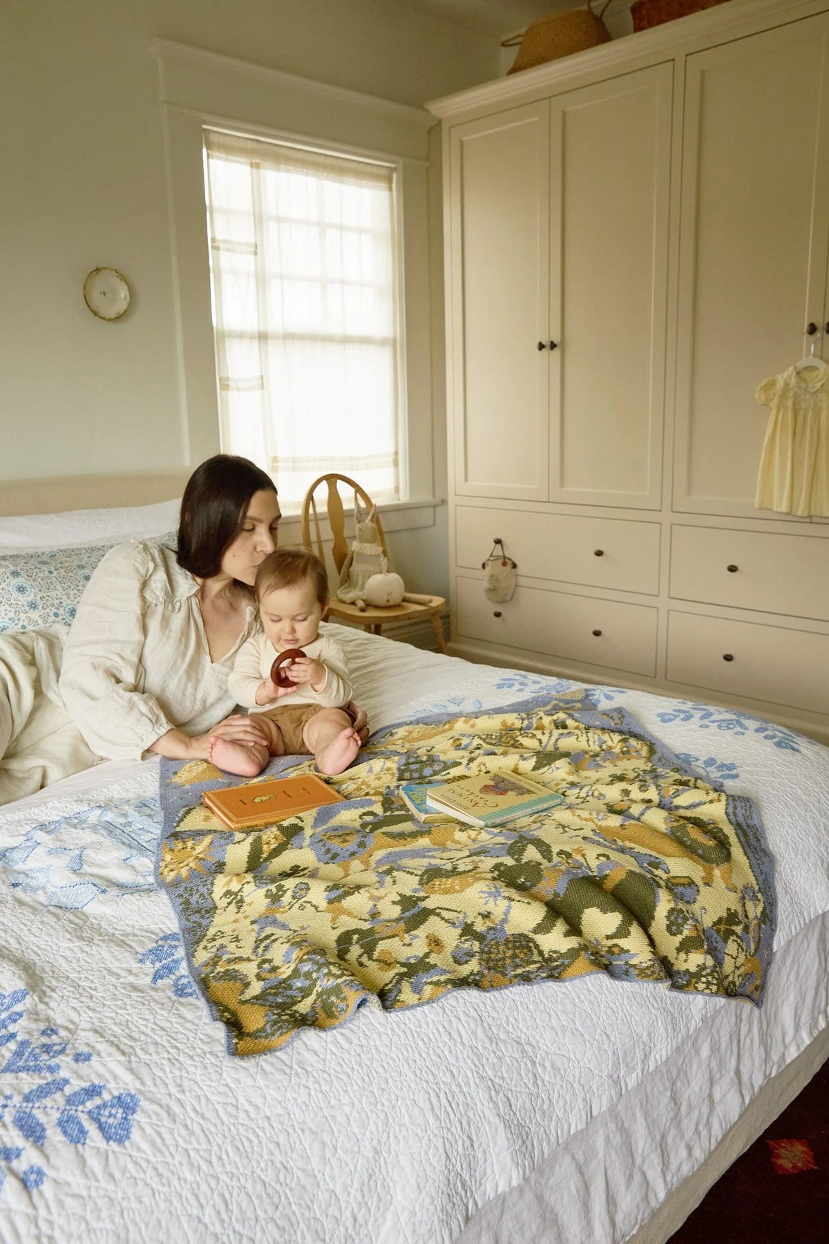 Adult and child seated on a bed beside the French Cirque Sky Blue blanket, with books on its circus-patterned knit.
