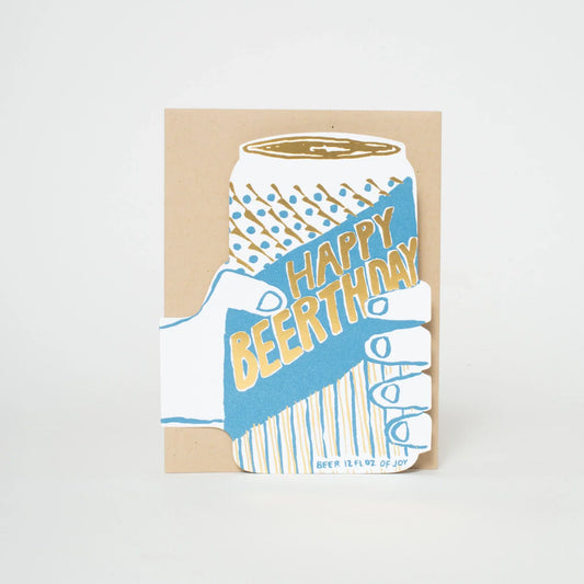 A square greeting card featuring a stylized illustration of a hand holding a beer can with the words 'Happy Beerthday' in gold foil on a blue background, set against a plain white studio backdrop.