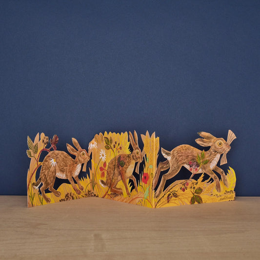 Hares Collage concertina card by Mark Hearld, fully extended showing three illustrated hares running through golden meadow grasses and wildflowers against a navy background