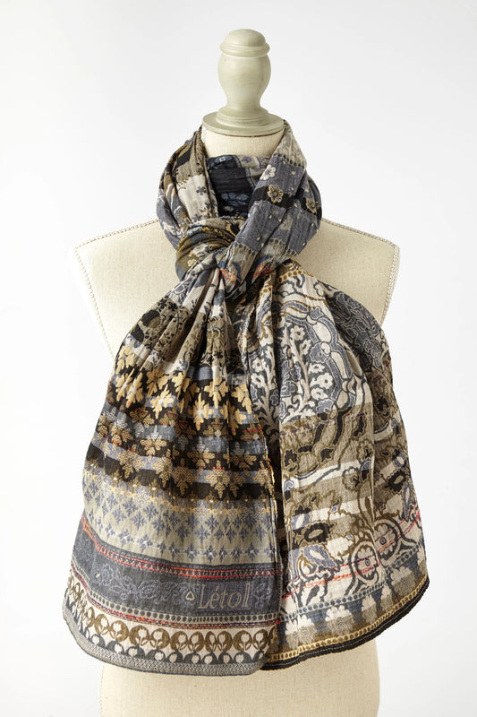 Létol Anoushka Scarf in Noir, draped and knotted on a linen dress form against a light grey background, showing a reversible Jacquard-woven organic cotton scarf with rich horizontal bands of pattern — one side featuring large oatmeal and gold paisley medallions and floral scrollwork on a black ground, the other showing intricate chambray and indigo botanical motifs on an oatmeal ground — with the woven Létol label visible at the lower center and a red accent selvedge at the hem
