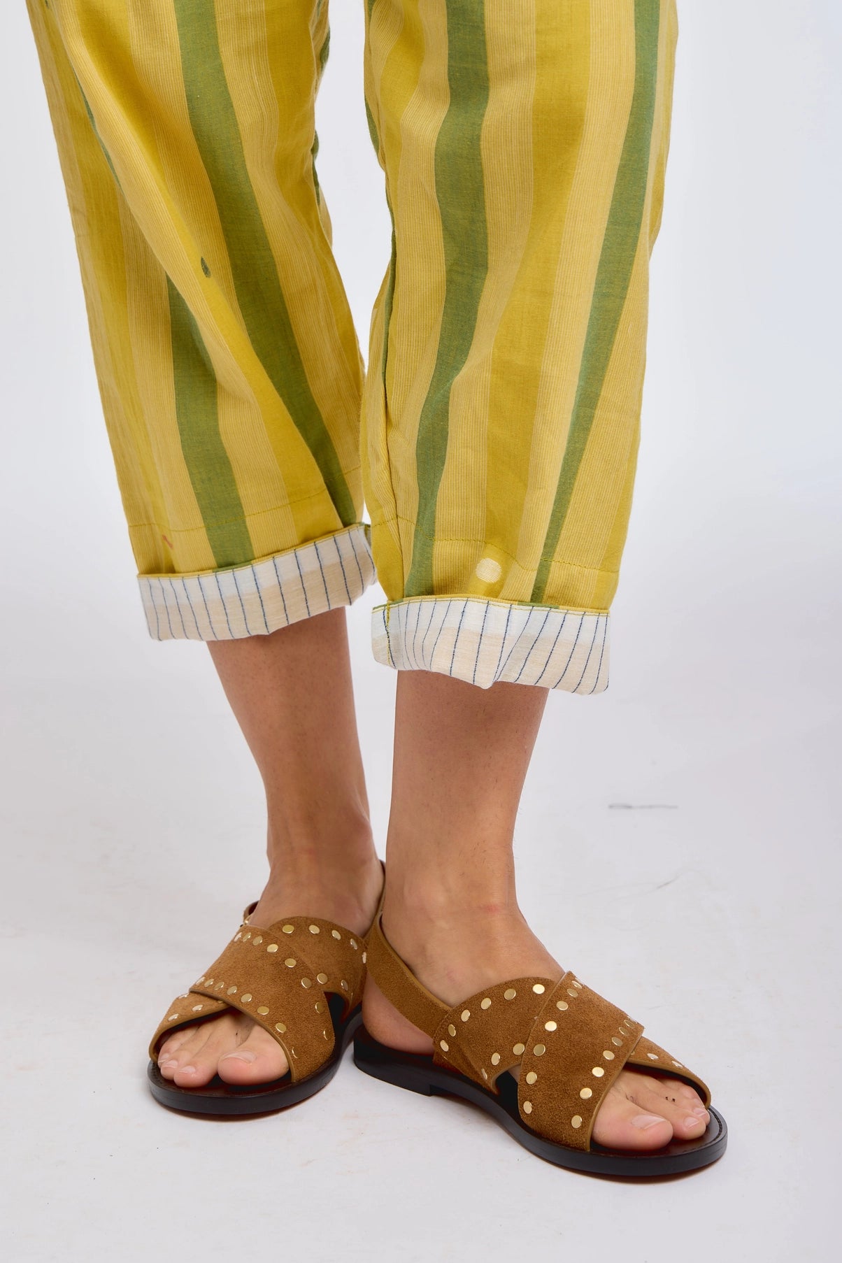 Close-up of DVAA Celeste Pant in Goldenrod hem and footwear detail against white studio backdrop showing goldenrod yellow and sage green wide vertical stripe handwoven cotton, cropped tapered leg with contrast white pinstripe cuff turn-up, styled with tan leather gold-studded crossband slide sandals
