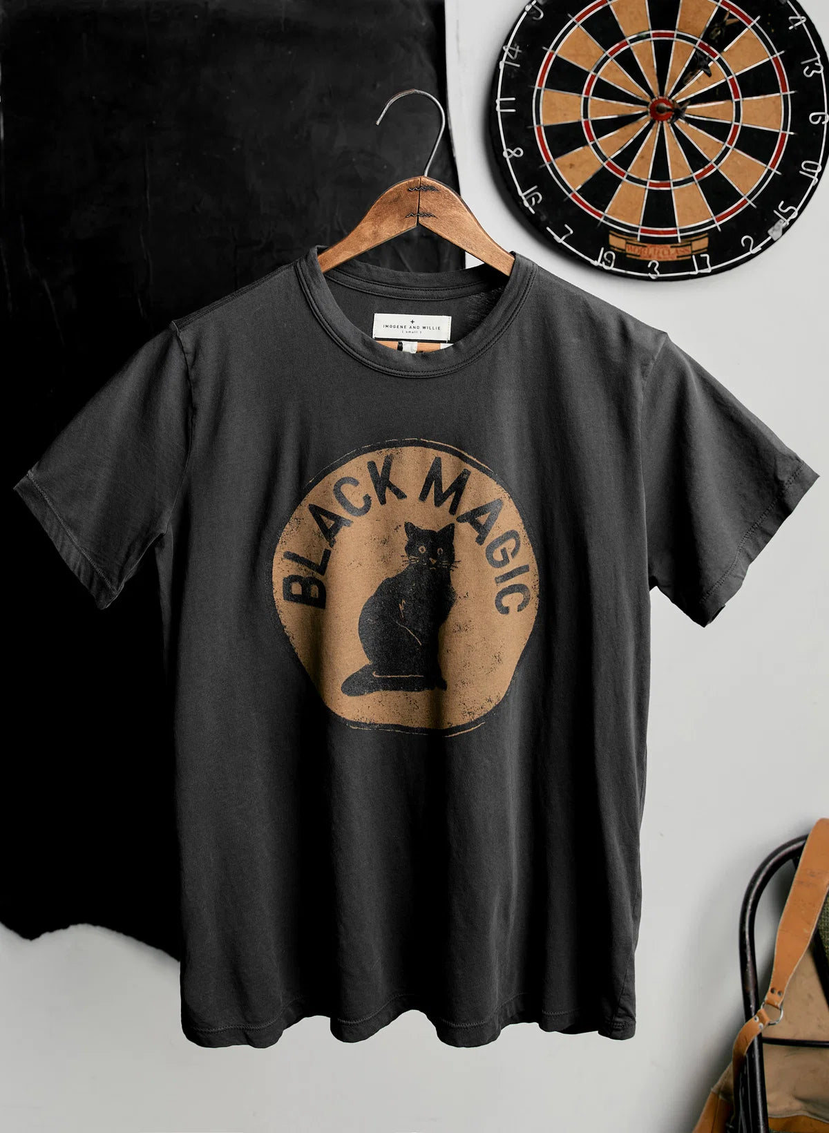Faded black short-sleeve tee on a wooden hanger with a vintage-style screen print of a black cat inside a circle reading "Black Magic," styled in front of a dartboard.
