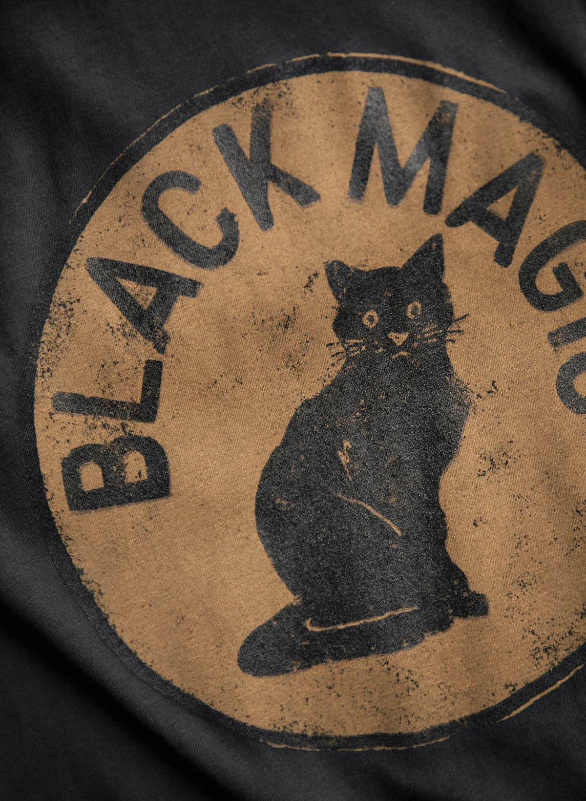 Close-up of the distressed "Black Magic" screen print on faded black cotton, featuring a black cat illustration in a tan circle with aged texture.
