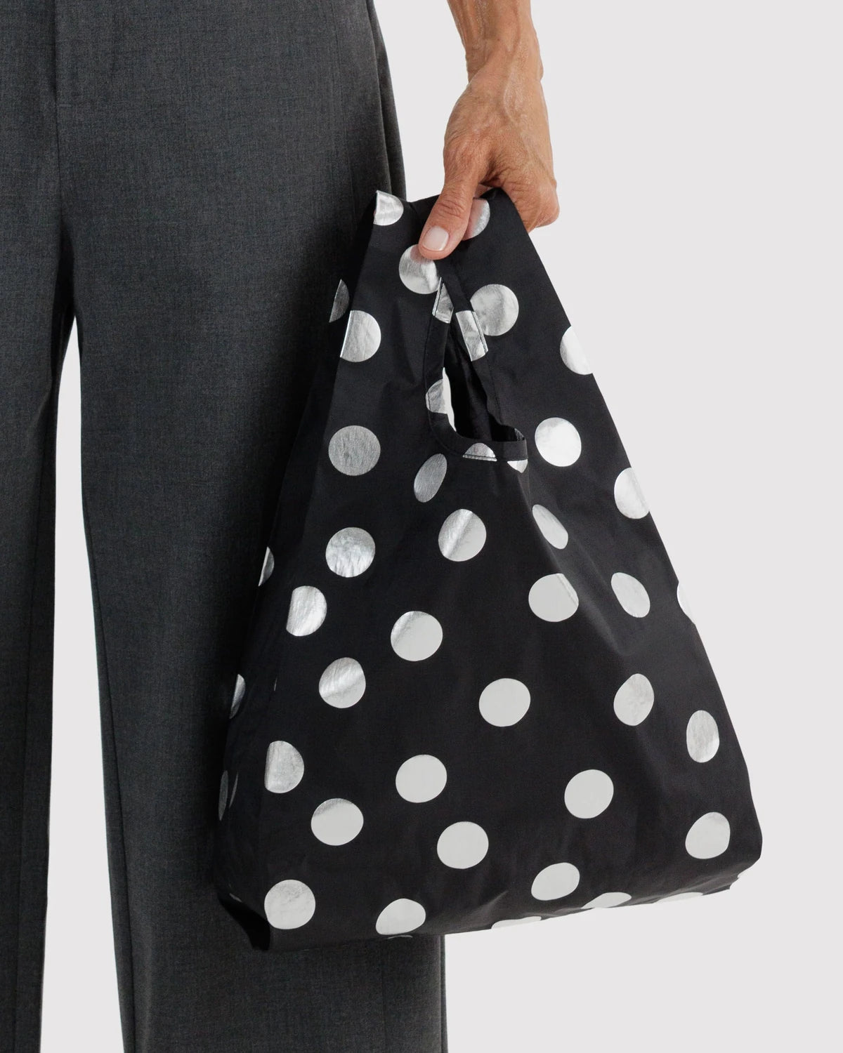 BAGGU Baby Baggu in Metallic Silver Polka Dot, held at a model’s side to show the small bag’s size and shiny silver dots on black fabric.
