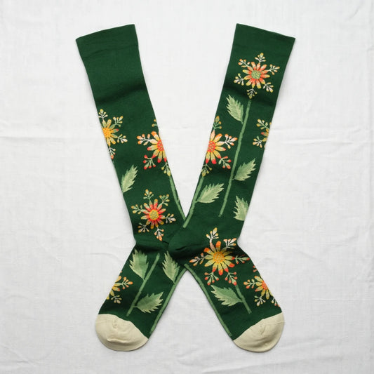 Bonne Maison Forest Snowflake Knee High Socks, flat lay pair showing forest green body with scattered snowflake-like botanical flower print in yellow, orange, and red with cream heel and toe
