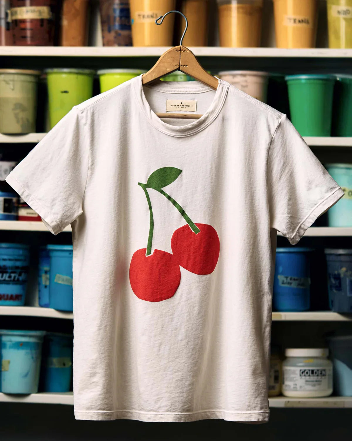 The Cherry Tee by Imogene + Willie on a wooden hanger in front of shelves of colorful paint cans, showing the full front with a large screen-printed cherry graphic — two red cherries with a green stem and leaf — on a white cotton tee