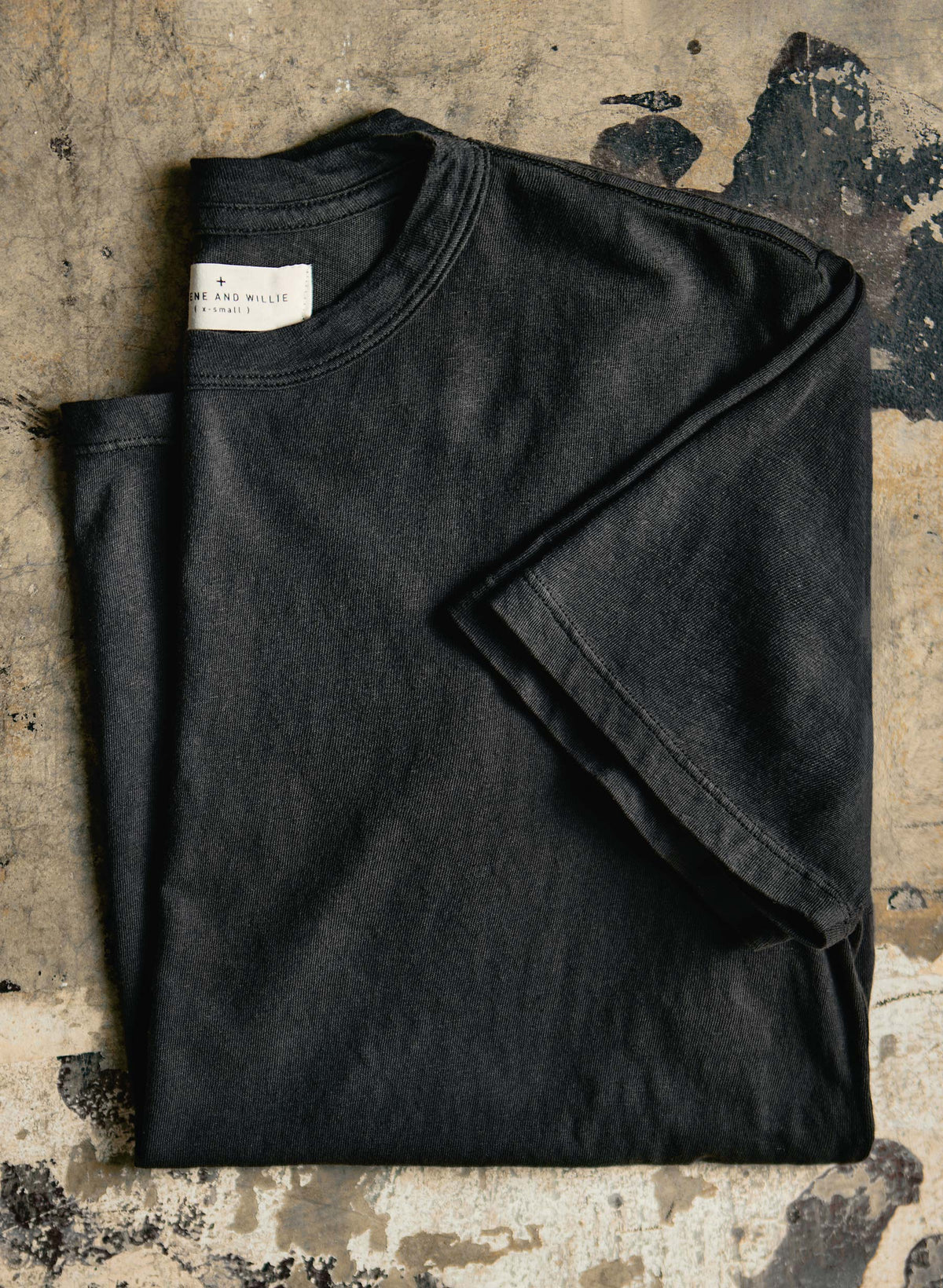 Imogene + Willie Classic Knit Crew in Black, folded t-shirt with label visible, shown on a worn concrete surface — detail view
