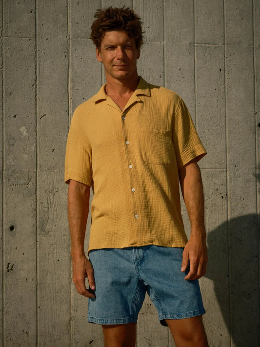 Model wearing Mollusk Surf Shop Cosmo Shirt in Saffron, full front view showing warm saffron yellow 100% cotton double gauze, open camp collar, short sleeves, 5-button placket with natural corozo-style buttons, and single left-chest patch pocket, styled with blue denim shorts against a concrete wall
