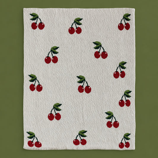Flat lay of the Dotter Cotton Knit Baby Blanket with Cherries, showing the cream knit face with an allover intarsia pattern of red cherries with green stems and leaves, on a sage green background

