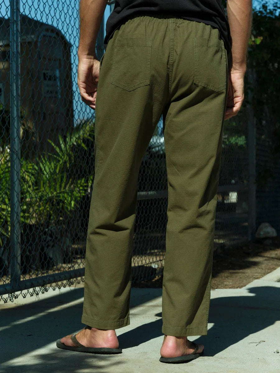 Model wearing Mollusk Surf Shop Drawstring Salvador Pants in Mash Green Ripstop, full back view showing olive green double-washed cotton ripstop, roomy straight-leg silhouette, elastic drawstring waistband, two back patch pockets, and straight hem with side vents, styled with black t-shirt and olive flip flops near chain-link fence
