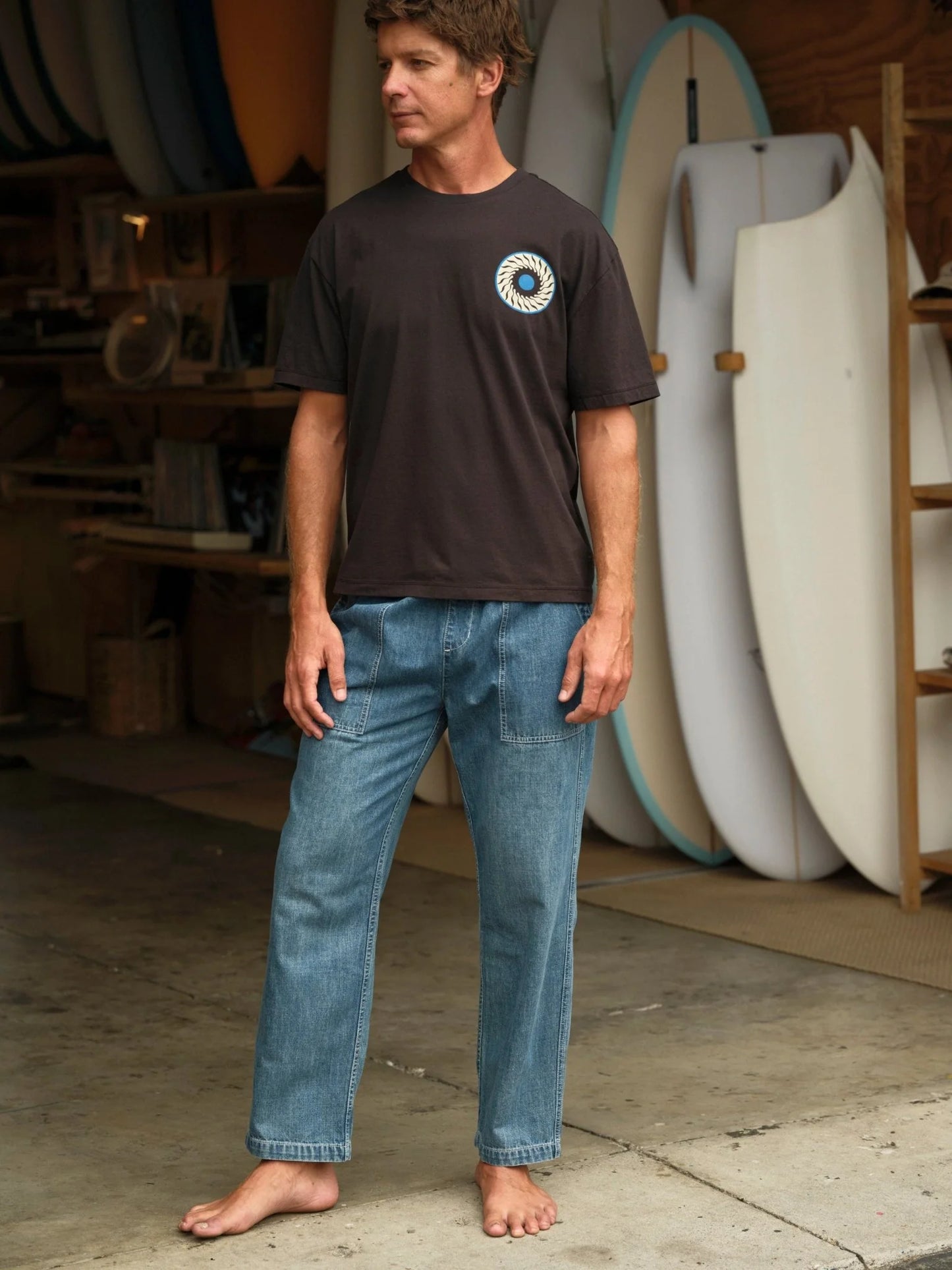 Model wearing Mollusk Surf Shop Drawstring Salvador Pants in Vintage Indigo Denim, full front view standing barefoot in a surf shaping room with surfboards in background, showing vintage indigo lightweight denim, roomy straight-leg silhouette, front patch pockets, and carpenter panel seam detail, styled with dark brown Mollusk graphic t-shirt
