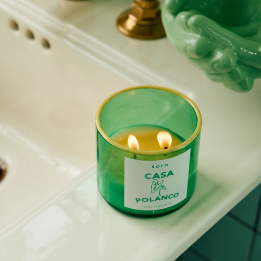 A lit Roen Casa Polanco candle in a green glass jar with a gold rim, displayed on a white surface against a green-tiled wall