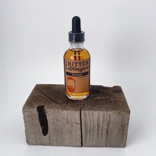 The Bitter Ginger Orange Bitters 2 oz glass dropper bottle with kraft paper label, displayed upright on a rustic weathered wood block against a white background