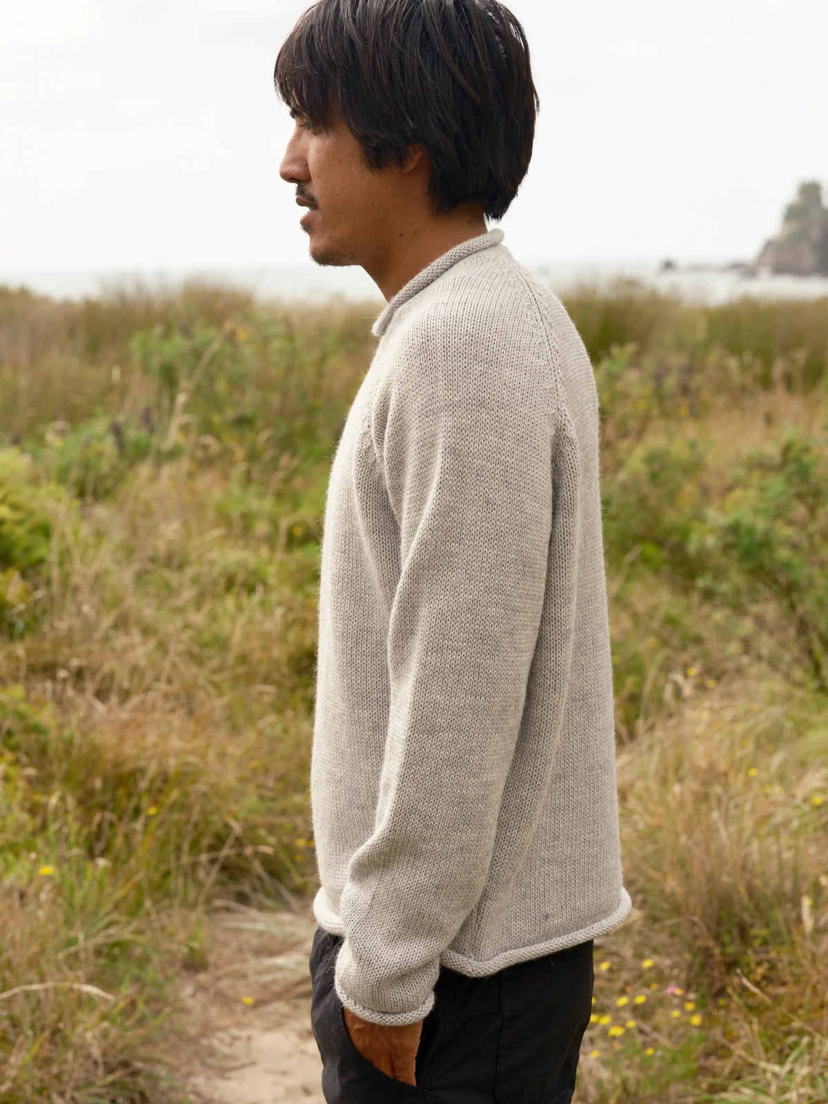 Pure side profile of a man standing on a coastal meadow path wearing the Mollusk Fisherman Sweater in Pearl Gray, showing the relaxed silhouette, raglan sleeve construction, rolled cuffs, and heathered light gray alpaca blend knit, with dry grasses and a coastal rock formation in the background

